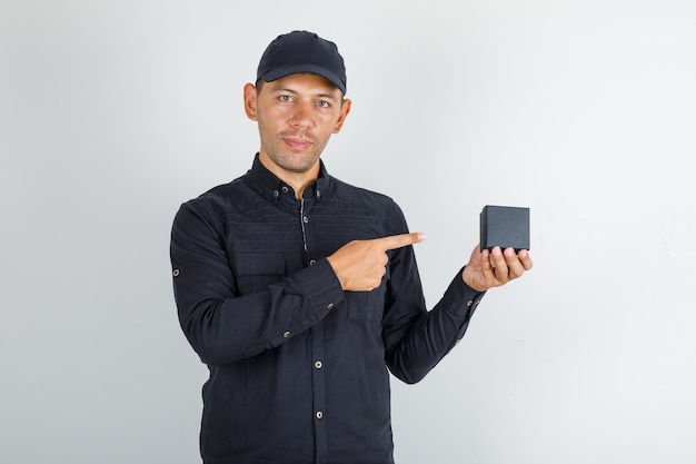 Joven apuntando con el dedo a la caja del reloj en camisa negra con gorra