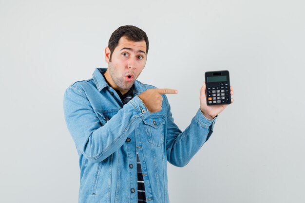 Joven apuntando con el dedo a la calculadora en camiseta, chaqueta y mirando sorprendido. vista frontal.