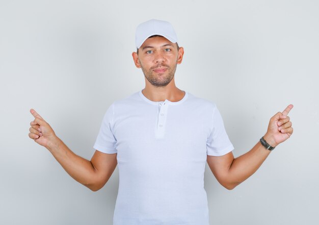 Joven apuntando con el dedo en camiseta blanca, gorra y mirando confiado, vista frontal