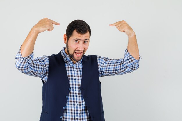 Foto gratuita joven apuntando con el dedo a su cabeza en camisa, chaleco y mirando confiado. vista frontal.