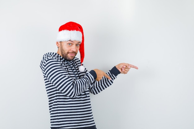 Joven apuntando a la derecha en sudadera con capucha, sombrero de Santa Claus y mirando alegre. vista frontal.