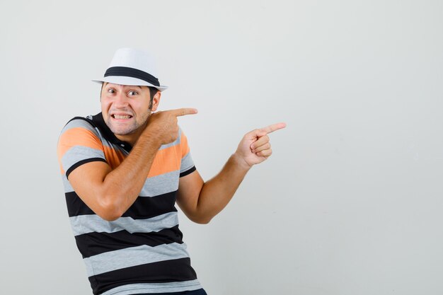 Joven apuntando a un lado con camiseta a rayas, sombrero y mirando loco espacio para texto