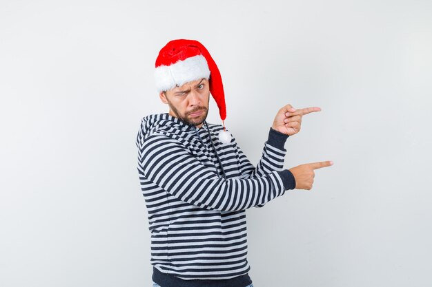 Joven apuntando hacia el lado derecho mientras parpadea en sudadera con capucha, gorro de Papá Noel y parece confundido,