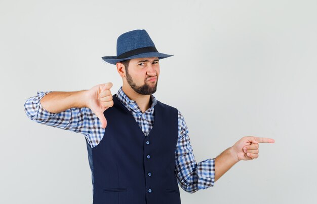 Joven apuntando hacia un lado, mostrando el pulgar hacia abajo en camisa, chaleco, sombrero y mirando descontento.