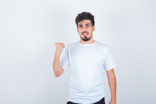 Joven apuntando a un lado con el pulgar en camiseta blanca y mirando confiado