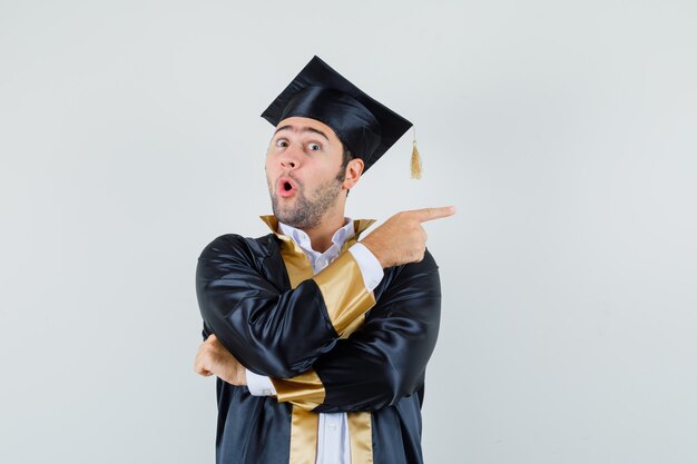 Joven apuntando hacia un lado en uniforme de posgrado y mirando sorprendido. vista frontal.