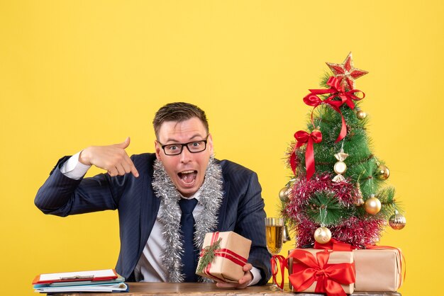 joven apuntando a sí mismo sentado en la mesa cerca del árbol de Navidad y presenta en amarillo