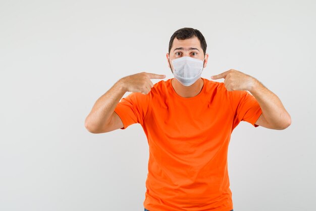 Joven apuntando a su máscara médica en camiseta naranja, vista frontal.