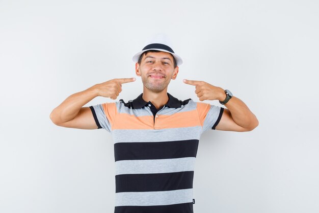 Joven apuntando a su sonrisa en camiseta y sombrero y mirando alegre