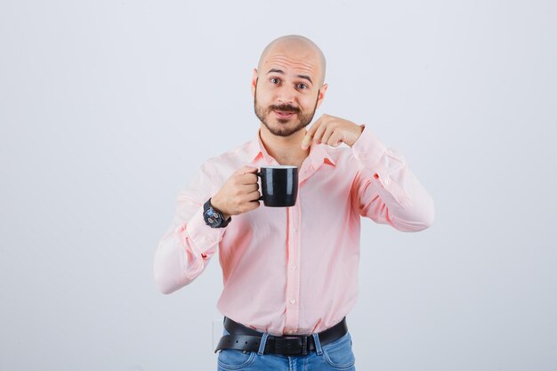 Joven apuntando a la taza en camisa rosa, jeans y mirando alarmado, vista frontal.