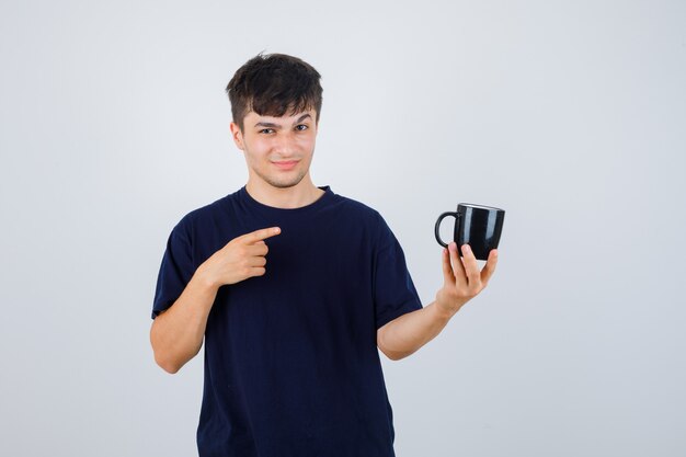 Joven apuntando a la taza en camiseta negra y mirando indeciso, vista frontal.