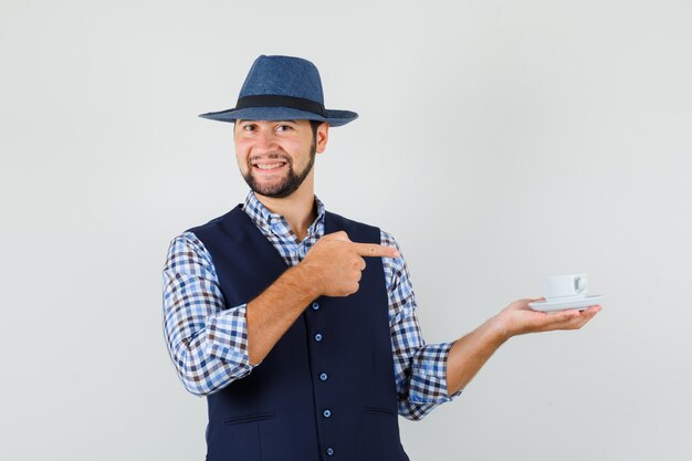 Joven apuntando a una taza de té en camisa, chaleco, sombrero y mirando alegre, vista frontal.