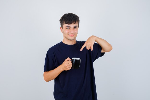 Joven apuntando a una taza de té en camiseta negra y mirando confiado, vista frontal.