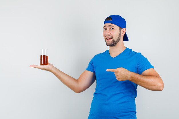 Joven apuntando a un vaso de bebida en camiseta azul y gorra y mirando feliz