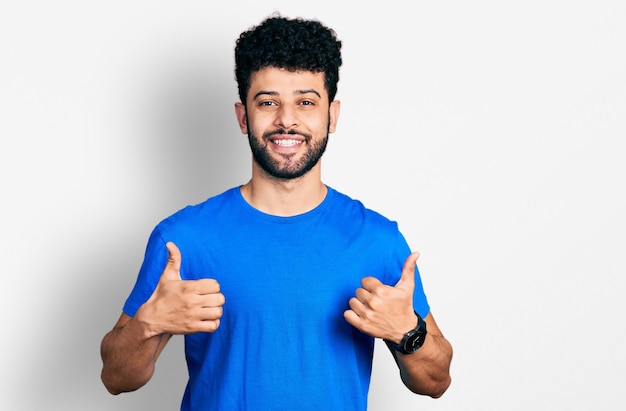 Joven árabe con barba con camiseta azul casual signo de éxito haciendo gesto positivo con la mano, pulgares arriba sonriendo y feliz. expresión alegre y gesto ganador.