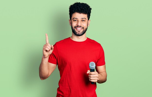 Joven árabe con barba cantando una canción usando un micrófono sonriendo con una idea o pregunta señalando con el dedo con cara feliz, número uno