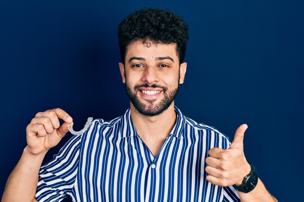 Joven árabe con barba sosteniendo alineador invisible ortodoncia y aparatos ortopédicos sonriendo feliz y positivo, pulgar arriba haciendo excelente y signo de aprobación