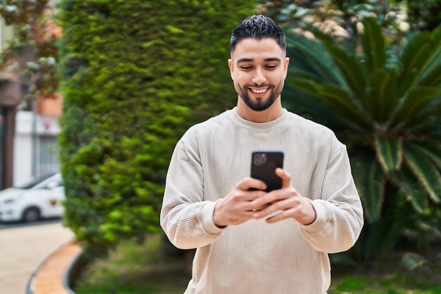 Joven árabe sonriendo confiado usando un teléfono inteligente en el parque