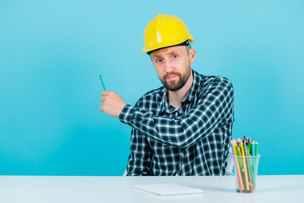 El joven arquitecto está apuntando a la pared trasera con un lápiz en la mano sobre fondo azul.