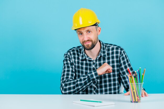 El joven arquitecto está mirando a la cámara sosteniendo el puño en el corazón sobre fondo azul.