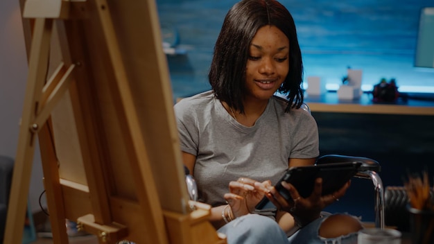 Foto gratuita joven artista negro con discapacidad mirando la tableta mientras está sentado en silla de ruedas en el taller. mujer afroamericana inválida que usa tecnología digital para inspirarse y dibujar obras maestras