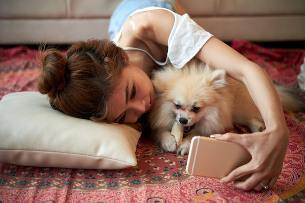 Joven asiática acostada en el piso con un pequeño perro mascota y tomar selfies con smartphone