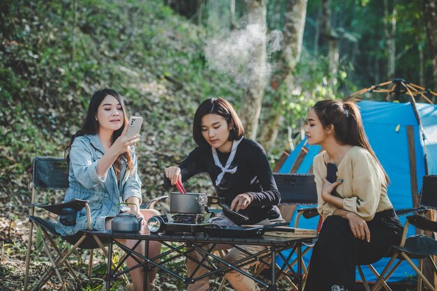 Una joven asiática cocinando y su amiga disfrutan de hacer la comida en una olla. Hablan y se ríen juntos mientras acampan en el parque natural.