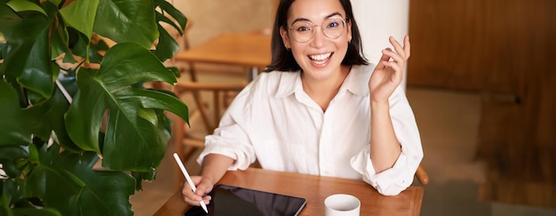 Foto gratuita joven asiática creativa sentada con una tableta digital dibujando con lápiz gráfico y garabateando sonriente