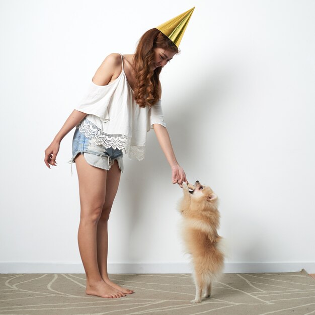 Joven asiática en divertido sombrero de fiesta jugando en casa con un pequeño perro mascota