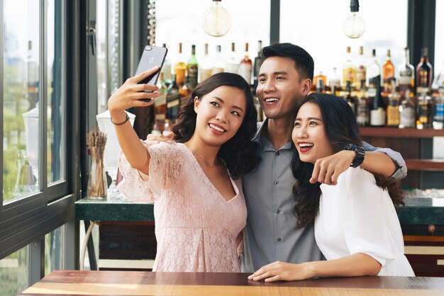 Joven asiática y dos mujeres abrazándose y tomando selfie en teléfono inteligente en el bar