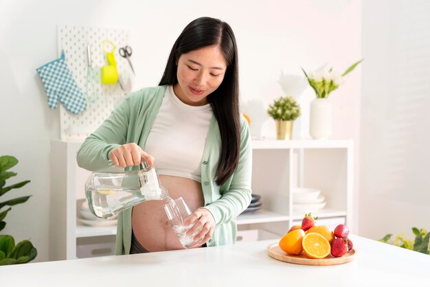 Joven asiática embarazada bebiendo agua en la cocina