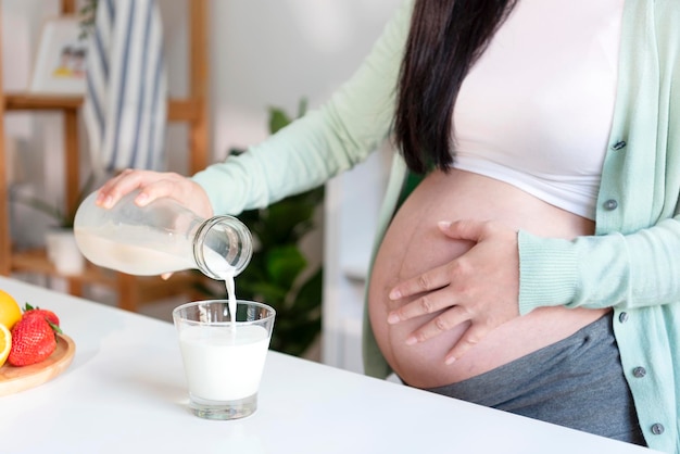 Joven asiática embarazada parada en la cocina sosteniendo un vaso de leche fresca