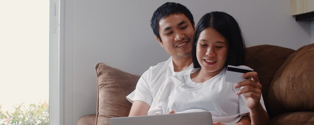 Joven asiática embarazada pareja de compras en línea en casa. Mamá y papá se sienten felices usando la tecnología portátil y la tarjeta de crédito comprando productos para bebés mientras están acostados en el sofá de la sala de estar en casa.