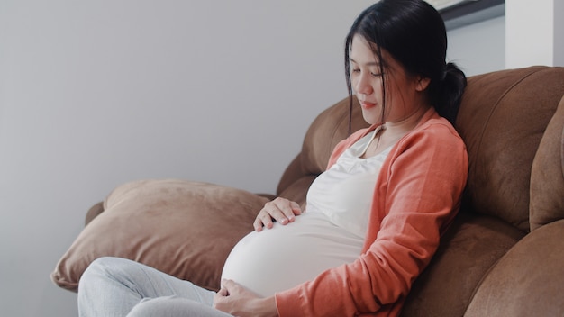 Joven asiática embarazada sosteniendo su vientre hablando con su hijo. Mamá se siente feliz sonriendo positiva y pacífica mientras cuida bebé, embarazo acostado en el sofá en la sala de estar en casa.