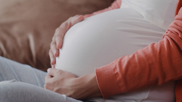 Joven asiática embarazada sosteniendo su vientre hablando con su hijo. Mamá se siente feliz sonriendo positiva y pacífica mientras cuida bebé, embarazo acostado en el sofá en la sala de estar en casa.