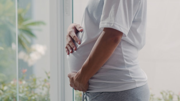 Joven asiática embarazada sosteniendo su vientre hablando con su hijo. Mamá se siente feliz sonriendo positiva y pacífica mientras cuida bebé, embarazo cerca de la ventana en la sala de estar en casa.