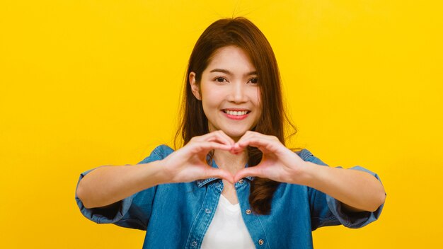 Joven asiática con expresión positiva, muestra el gesto de las manos en forma de corazón, vestido con ropa casual y mirando a la cámara sobre la pared amarilla. Feliz adorable mujer alegre disfruta el éxito.