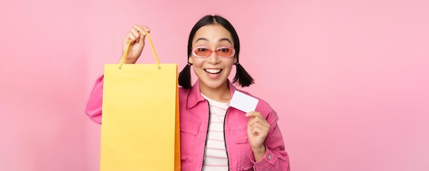 Una joven asiática feliz mostrando una tarjeta de crédito para comprar una bolsa comprando a la venta yendo a la tienda de pie sobre un fondo rosa