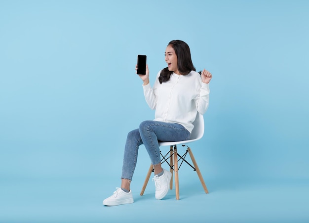 Una joven asiática feliz que muestra un teléfono inteligente de pantalla vacía mientras está sentada en una silla blanca