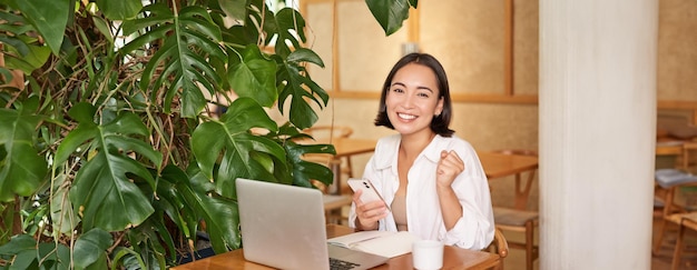 Foto gratuita una joven asiática feliz siente el éxito ganando y celebrando sentada con una computadora portátil y un teléfono inteligente en