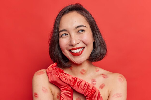 La joven asiática feliz y soñadora se concentra con una sonrisa dentuda, los hombros desnudos, tiene rastros de besos en el cuerpo, usa guantes aislados sobre un fondo rojo intenso. Emociones felices y concepto de belleza.