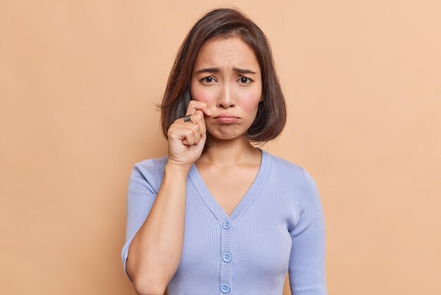 Una joven asiática frustrada e infeliz solloza y se frota las lágrimas se siente desesperada usa un jersey azul tiene una expresión insatisfecha calor roto aislado sobre una pared beige