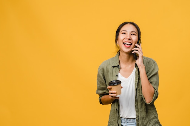 Joven asiática habla por teléfono y sostiene una taza de café con expresión positiva, sonríe ampliamente, vestida con un paño informal sintiendo felicidad y parada aislada en la pared amarilla. Concepto de expresión facial.
