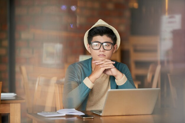 Joven asiática hipster hombre sentado en la cafetería con laptop y mirando