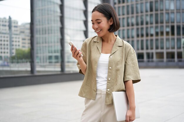 Joven asiática con laptop caminando por la calle y enviando mensajes de texto sonriendo mientras mira el smartph