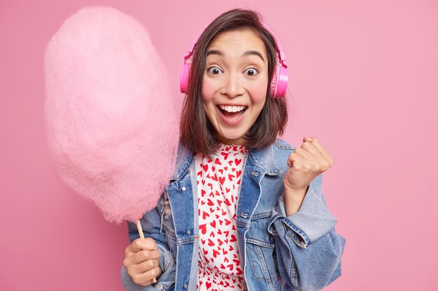 Una joven asiática llena de alegría escucha noticias asombrosas levanta el puño cerrado sonríe ampliamente celebra el triunfo come un delicioso hilo de azúcar endulzado escucha música a través de auriculares inalámbricos posa en el interior.