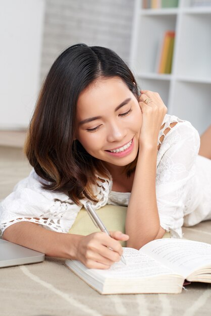 Joven asiática marcando texto en su libro acostado en el piso