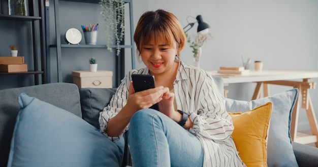 Joven asiática con mensaje de texto de teléfono inteligente o consultar las redes sociales en el sofá de la sala de estar de la casa