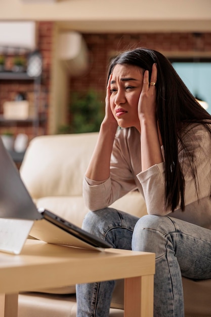 Joven asiática con migraña y dolor de cabeza mientras intenta trabajar desde casa. mujer cansada en el sofá que cubre la frente y los ojos rojos agotados infeliz enfermo malestar fiebre depresión