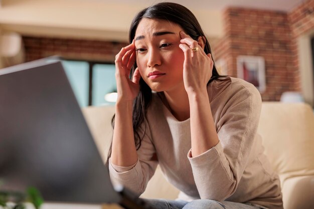 Joven asiática con migraña y dolor de cabeza mientras intenta trabajar desde casa. Mujer cansada en el sofá que cubre la frente y los ojos rojos agotados infeliz enfermo malestar fiebre depresión